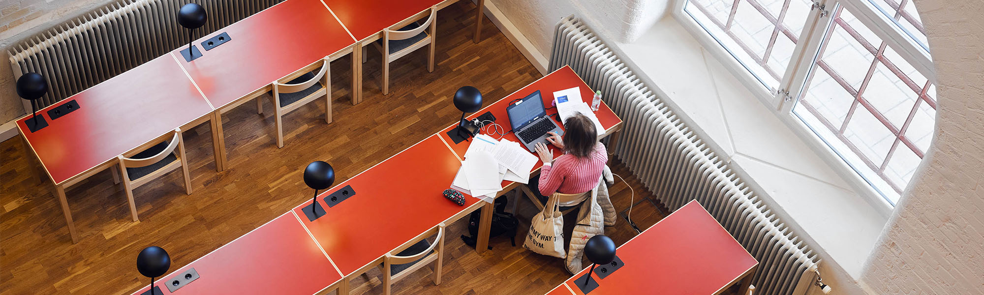 En ensam student sitter och studerar med en laptop och en massa papper utspridda på det röda bordet. Bordet är intill ett stort fönster. Bilden är tagen uppifrån.