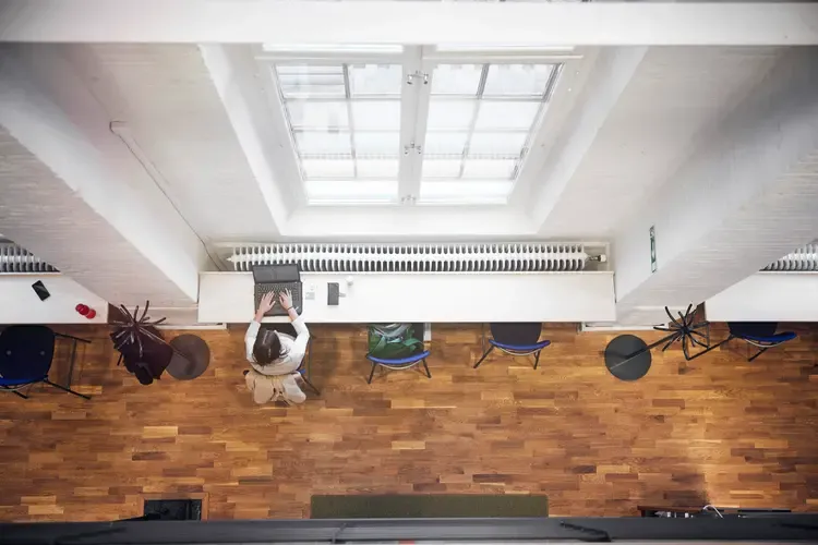 En bild tagen från ovan på ett bord intill ett högt fönster, där en student sitter och skriver på en dator.