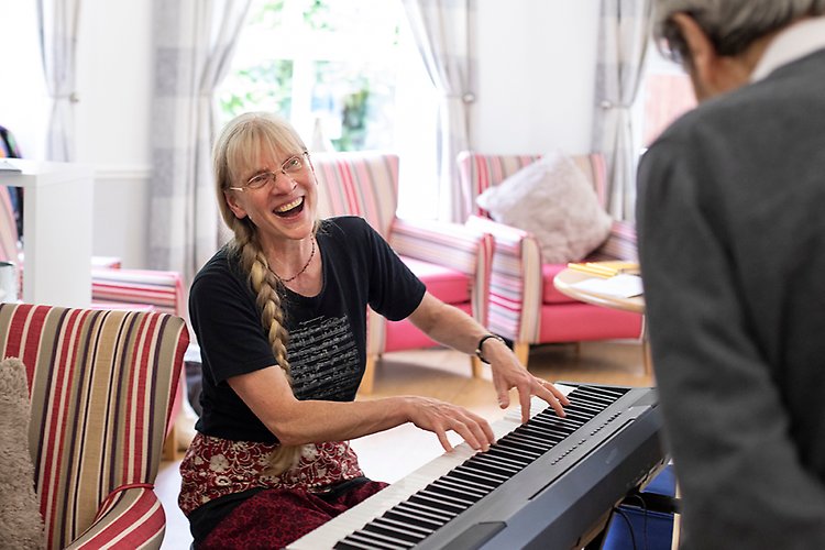 Kvinna sitter vid ett elpiano och spelar. I förgrunden står en äldre man. 